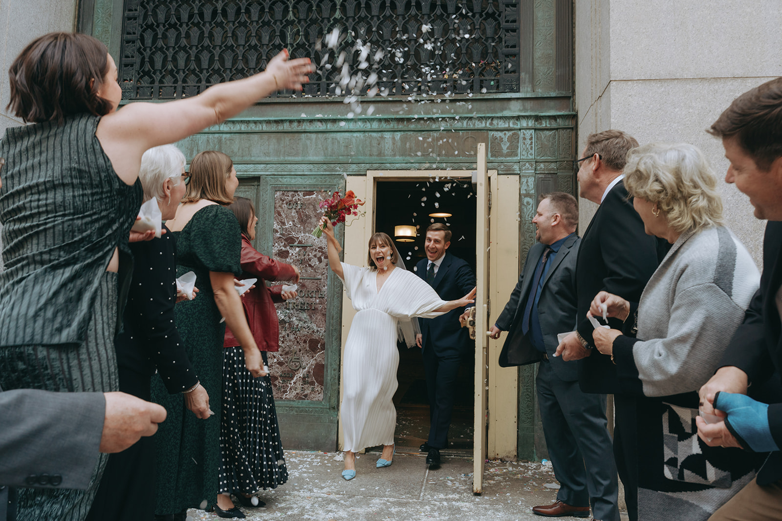 Rachel and Dave’s 2 day celebration Bride and groom walk through city hall doors to their friends and family throwing confetti by NYC city hall wedding photographer Elsie Goodman.