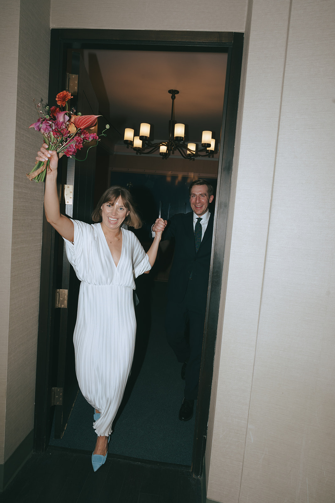 Rachel and Dave’s 2 day celebration Bride and groom walk through the doorway, she holds her bouquet above her head by NYC city hall wedding photographer Elsie Goodman.