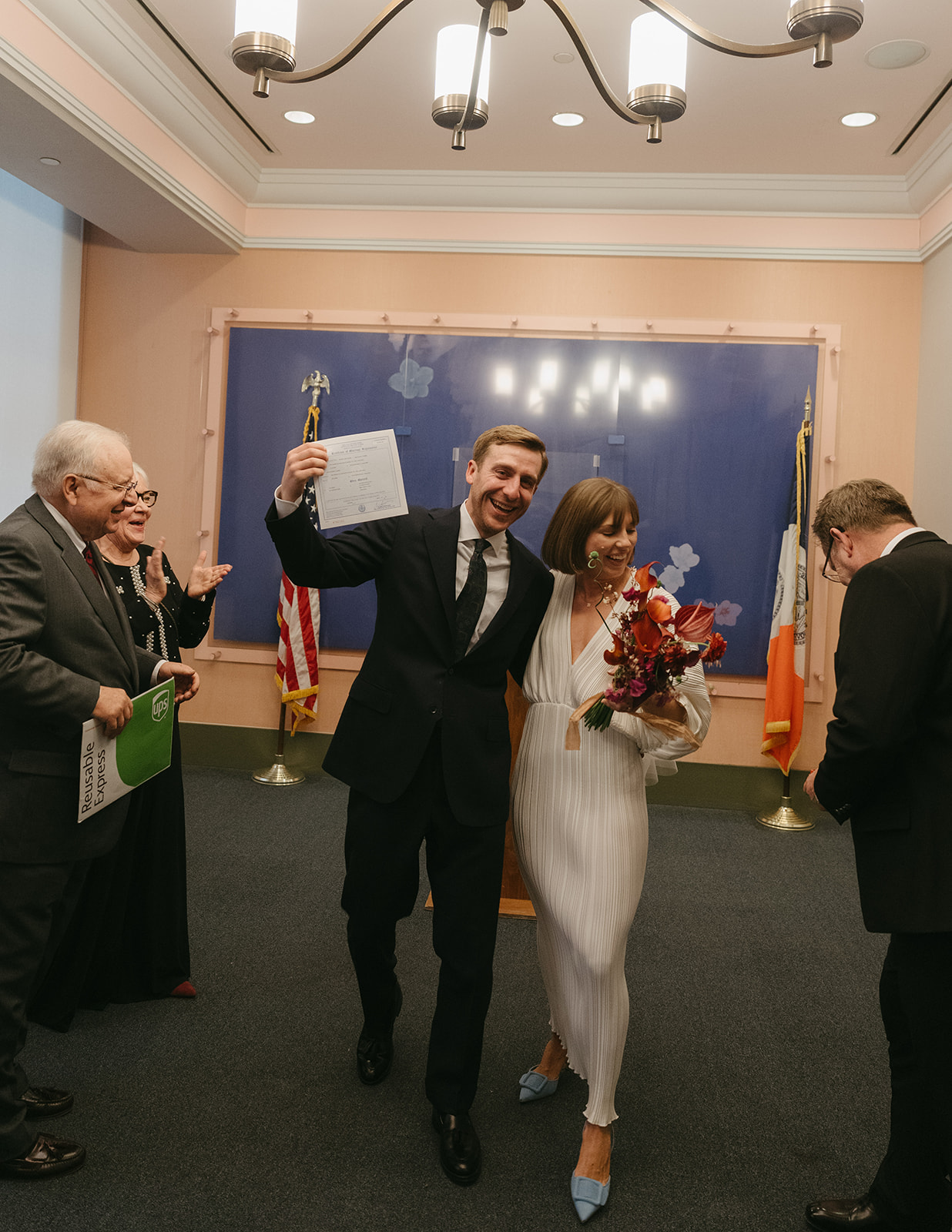 Rachel and Dave’s 2 day celebration Just married! Groom with his arm around his bride showing off their marriage certificate as they walk away from the marriage podium by NYC city hall wedding photographer Elsie Goodman.