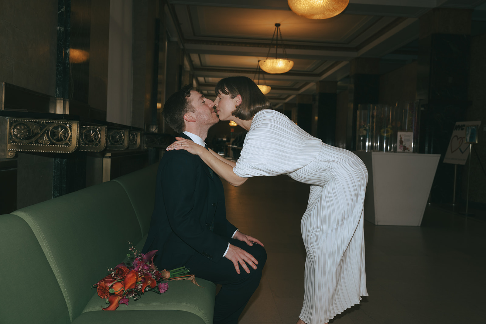 Rachel and Dave’s 2 day celebration Groom sits on a green bench, bride leans over, with her hands on his shoulders kissing him by NYC city hall wedding photographer Elsie Goodman.