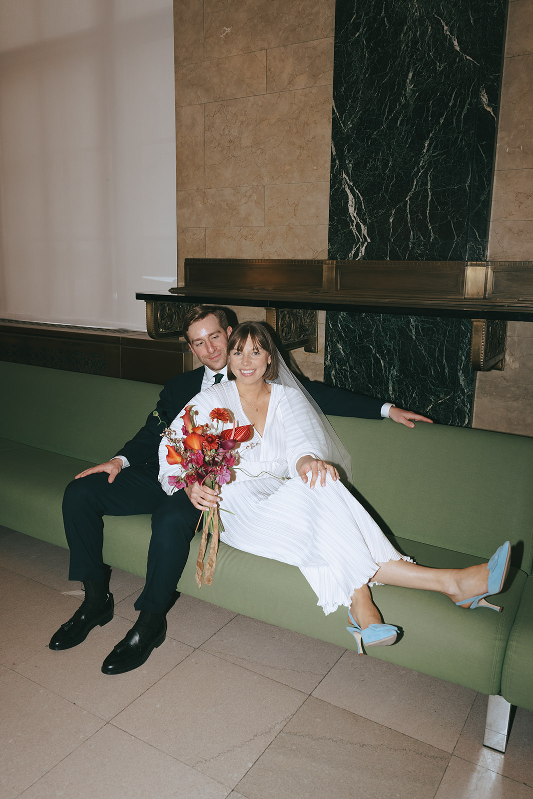 Rachel and Dave’s 2 day celebration Groom sits on a green couch looking at his bride, she is smiling at the camera with her feet up on the couch, showing off her light blue heels by NYC city hall wedding photographer Elsie Goodman.