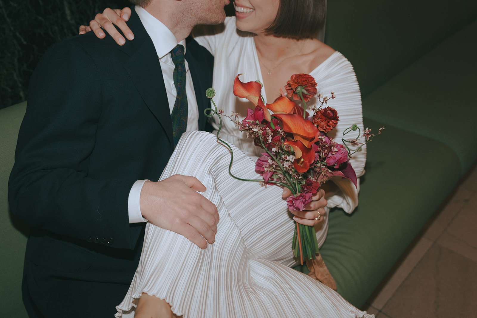 Bride sits with her legs in the groom's lap while sitting on a green couch, smiling, she holds her bouquet by NYC city hall wedding photographer Elsie Goodman.