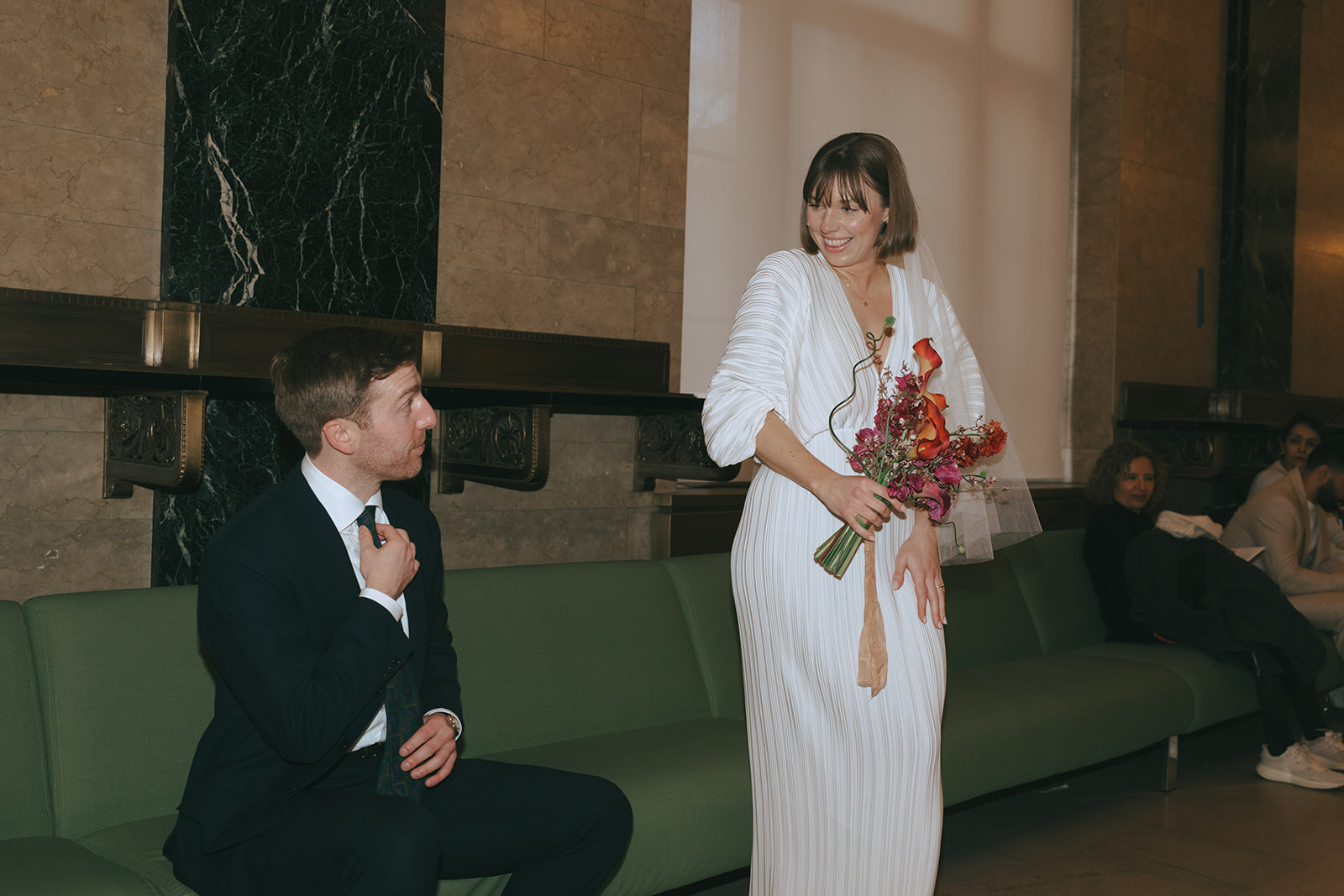 Bride stands with her bouquet looking at the groom as he sits and adjusts his tie, they are at NY City Hall by NYC elopement photographer Elsie Goodman.