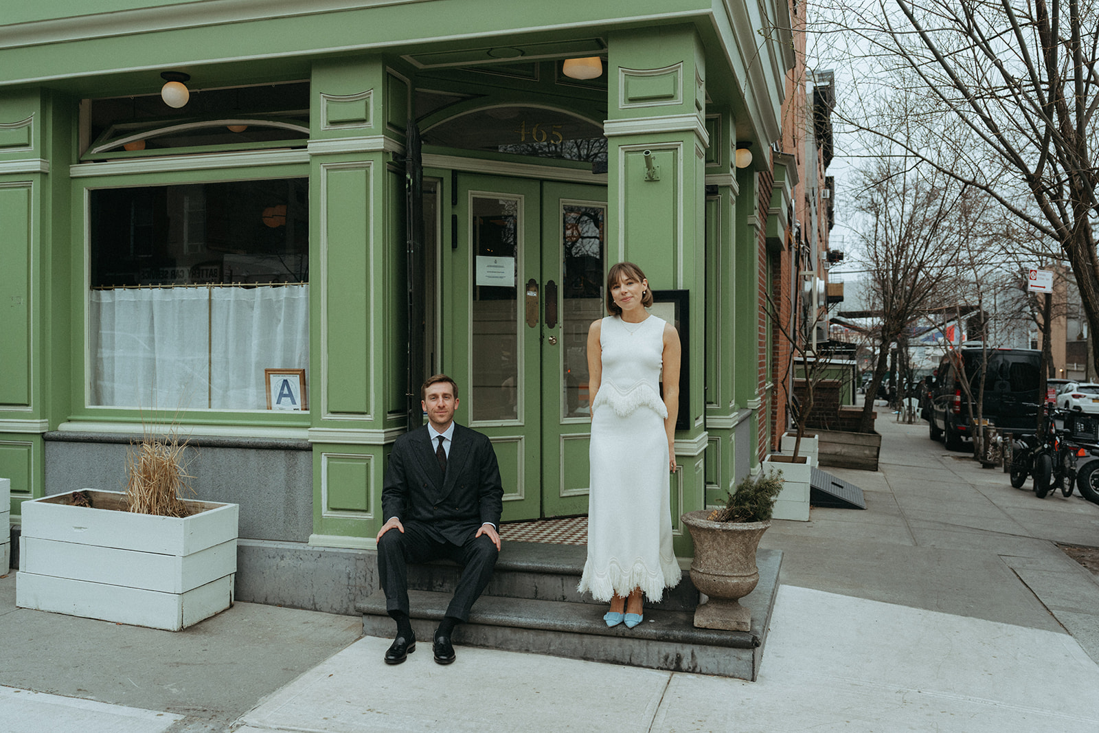 Rachel and Dave’s 2 day celebration Bride stands on the step, groom sits in front of Franks restaurant NYC by NYC elopement photographer Elsie Goodman.