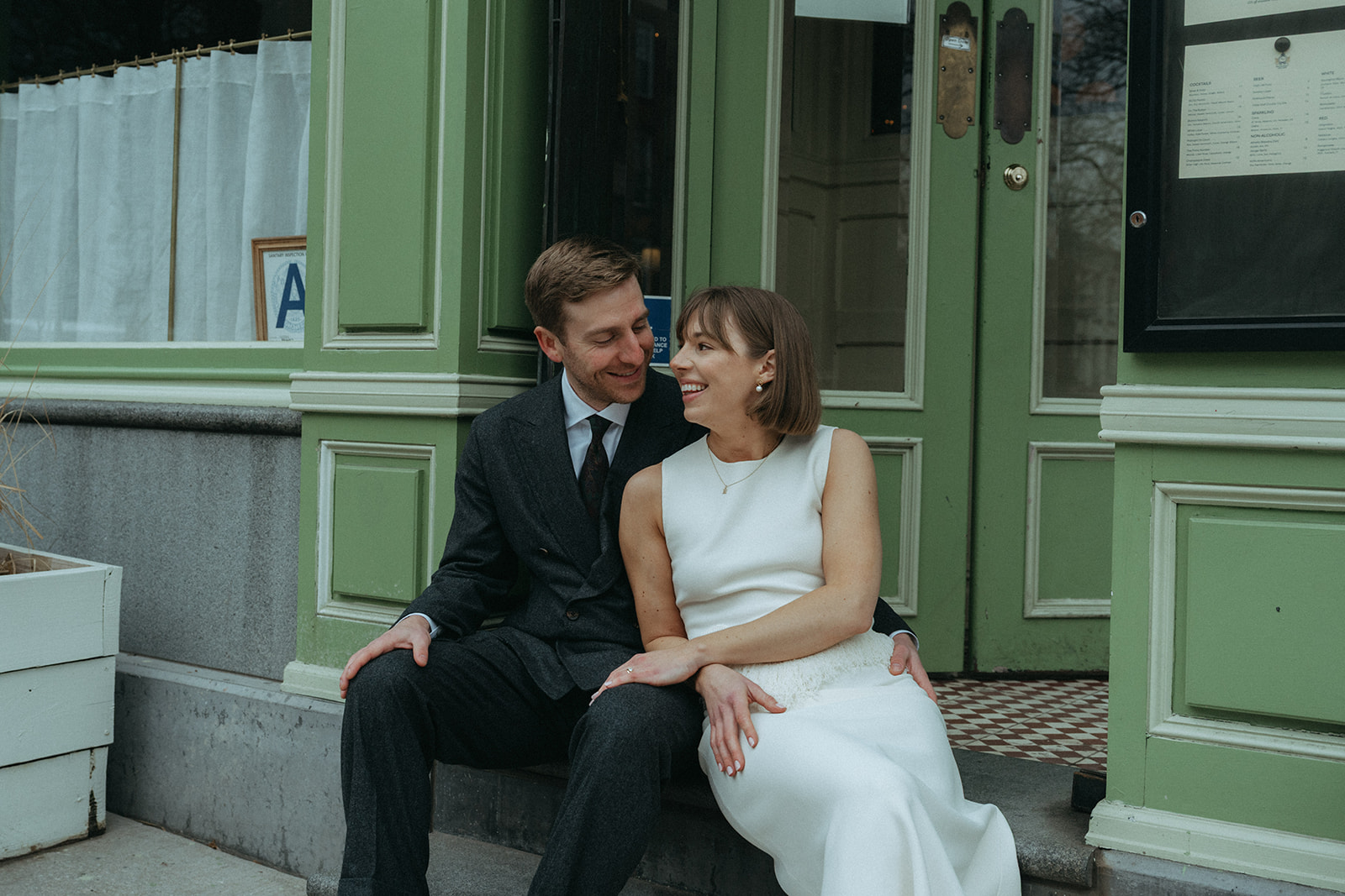 Rachel and Dave’s 2 day celebration Bride and groom sit on the step in front of franks leaning into each other, smiling, by NYC elopement photographer Elsie Goodman.