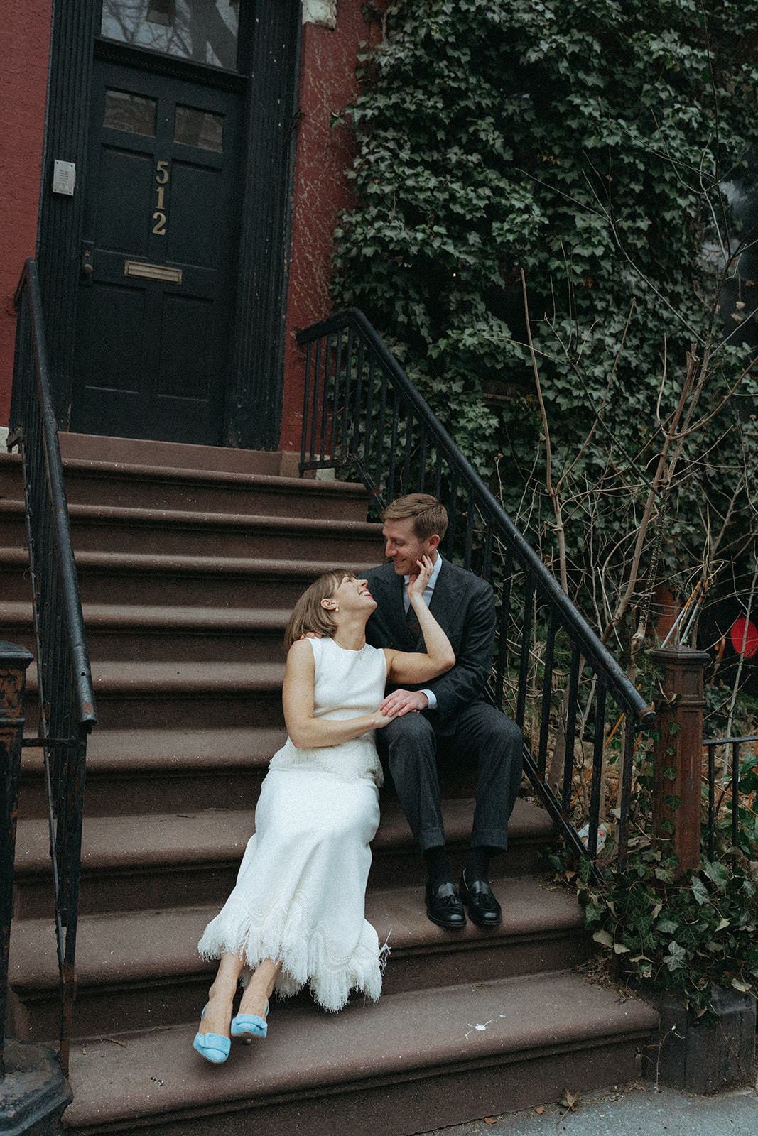 Rachel and Dave’s 2 day celebration Bride in a vintage wedding dress and blue suede shoes sits on a stoop looking up to her groom by NYC elopement photographer Elsie Goodman.