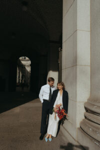 Bride wearing the grooms jacket leaning against a building as he looks at her by NYC city hall wedding photographer Elsie Goodman.