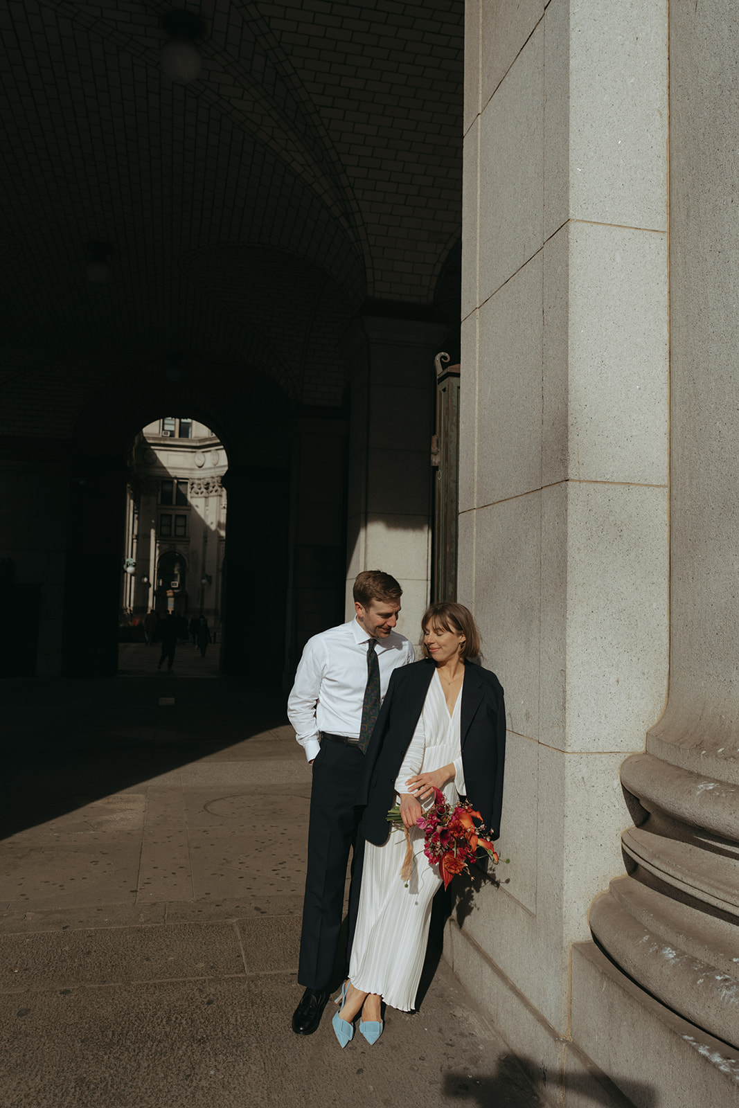 Rachel and Dave’s 2 day celebration Bride wearing the grooms jacket leaning against a building as he looks at her by NYC city hall wedding photographer Elsie Goodman.