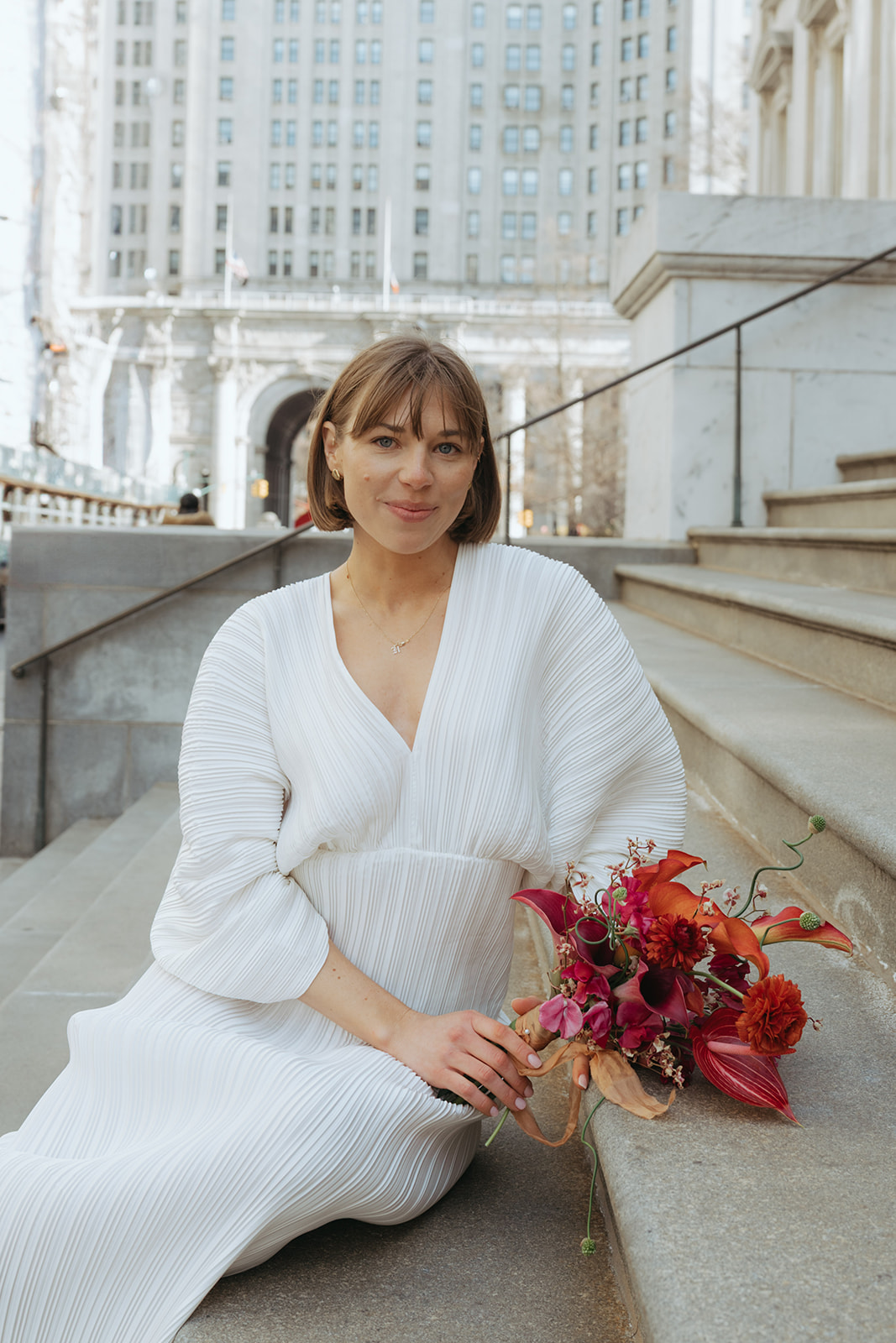 Rachel and Dave’s 2 day celebration Portrait of a bride in a vintage wedding dress, holding her bouquet, sitting on the steps of city hall by NYC city hall wedding photographer Elsie Goodman.