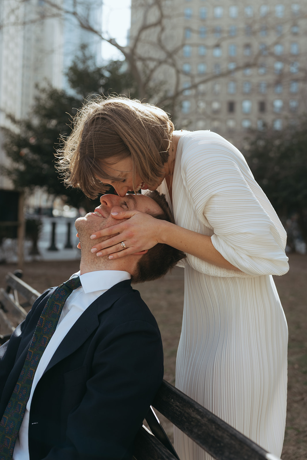 Rachel and Dave’s 2 day celebration Groom sits on a park bench, the bride in a white vintage dress is behind him kissing his forehead by NYC city hall wedding photographer Elsie Goodman.