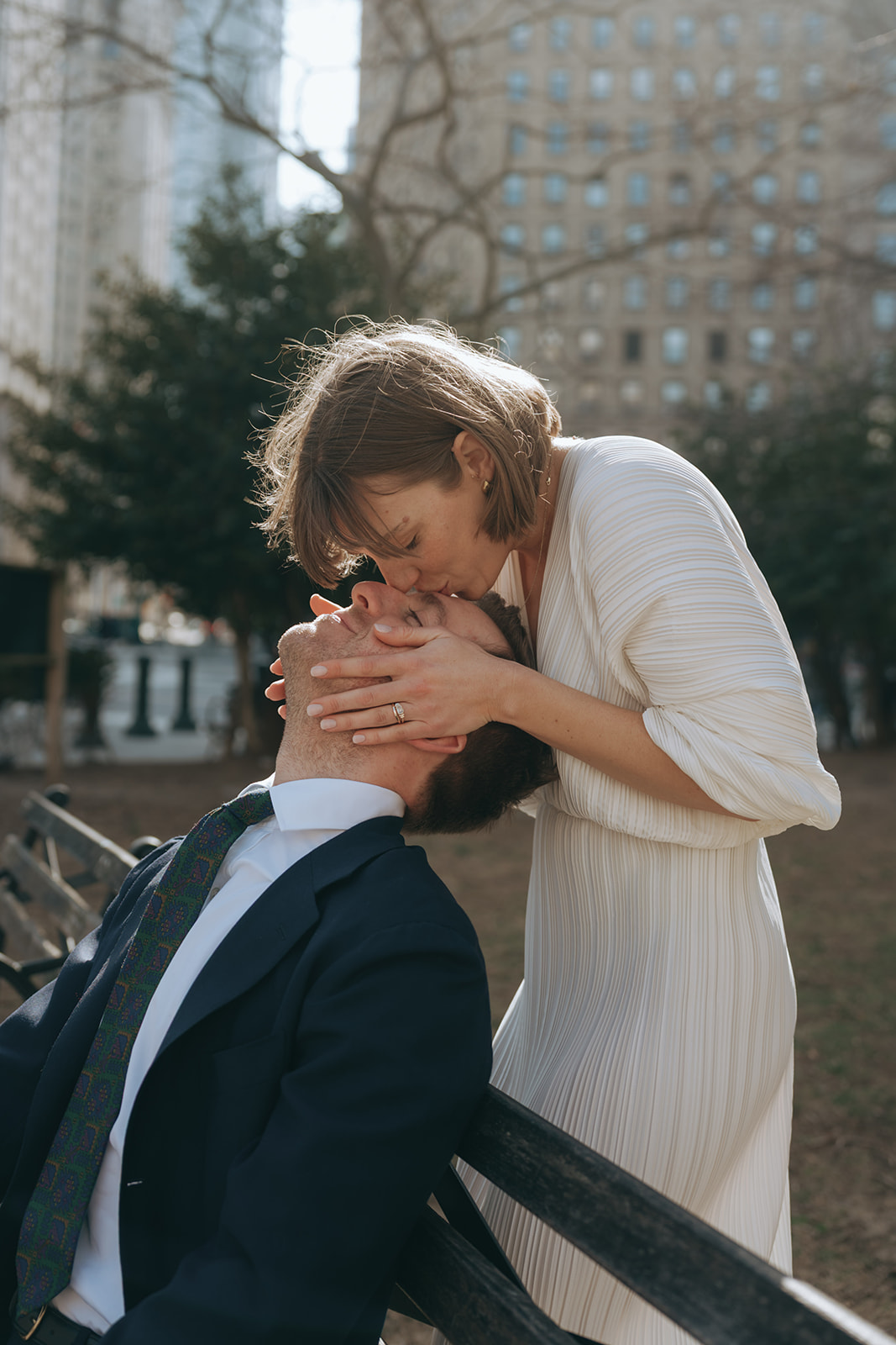 Rachel and Dave’s 2 day celebration Groom sits on a park bench, the bride in a white vintage dress is behind him kissing his forehead by NYC city hall wedding photographer Elsie Goodman.