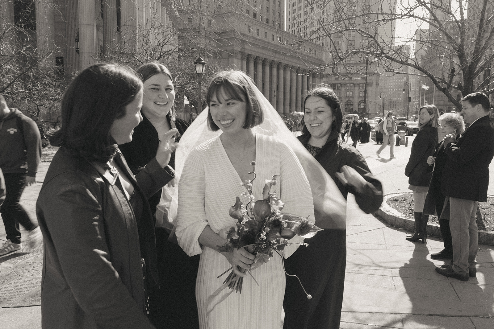 Rachel and Dave’s 2 day celebration Black and white photo of the bride in her vintage wedding dress, smiling outside of NY city hall with her friends and family, by NYC city hall wedding photographer Elsie Goodman.