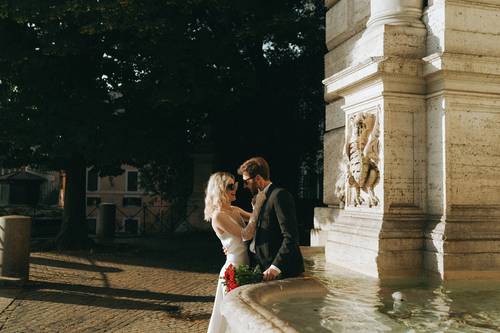 Bride in a white dress, gloves and sunglasses, groom in a tux leans again a fountain as she fixes his bowtie by Rome elopement photographer Elsie Goodman.