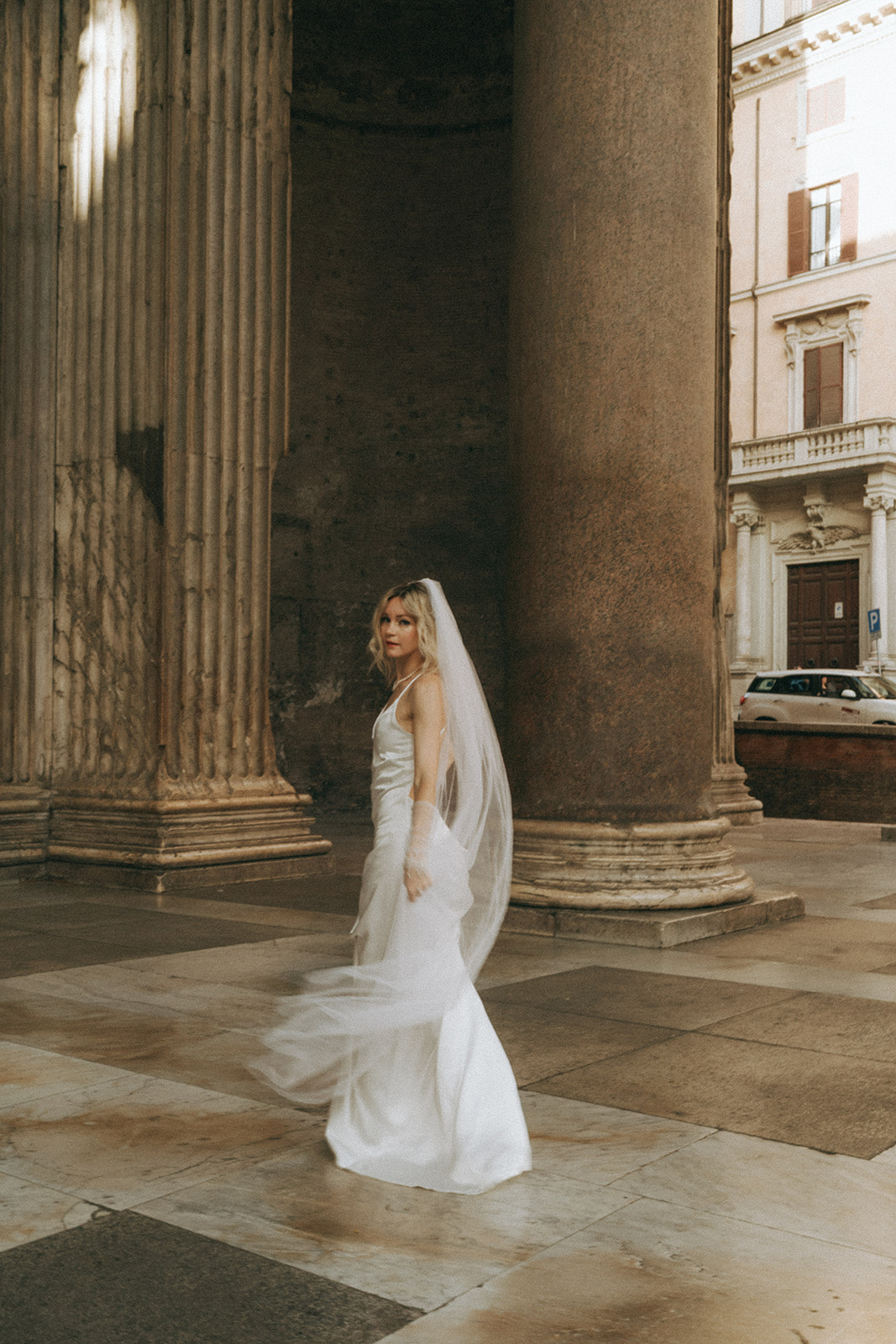 Full body shot of a bride in a full-length white dress and floor-length veil looking over her shoulder at the Pantheon by Rome elopement photographer Elsie Goodman.