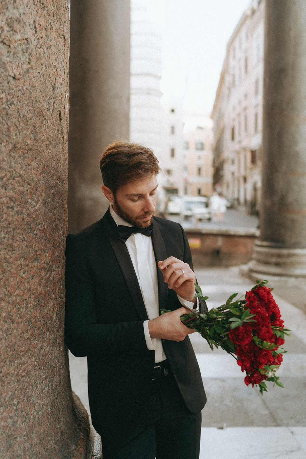 Groom in a tux leaning up against the columns of the Pantheon adjusting his cufflinks while holding a bouquet of roses by Rome elopement photographer Elsie Goodman.