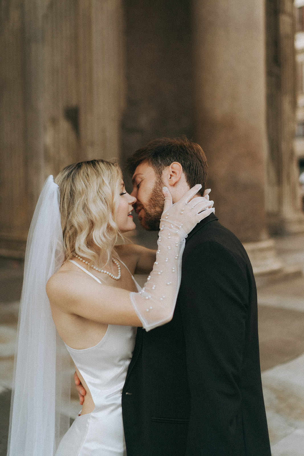 Bride and groom leaning into a kiss. She has on a long white dress, long veil and elbow length gloves, he is wearing a tux, by Rome elopement photographer Elsie Goodman.
