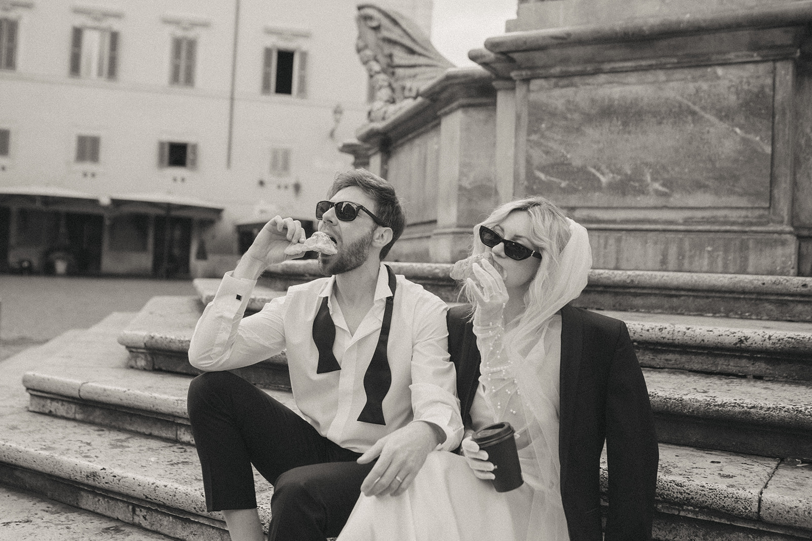 Bride and groom sitting on stone steps, both taking a bite out of their croissants, Trastevere coffee shop by Rome elopement photographer Elsie Goodman.