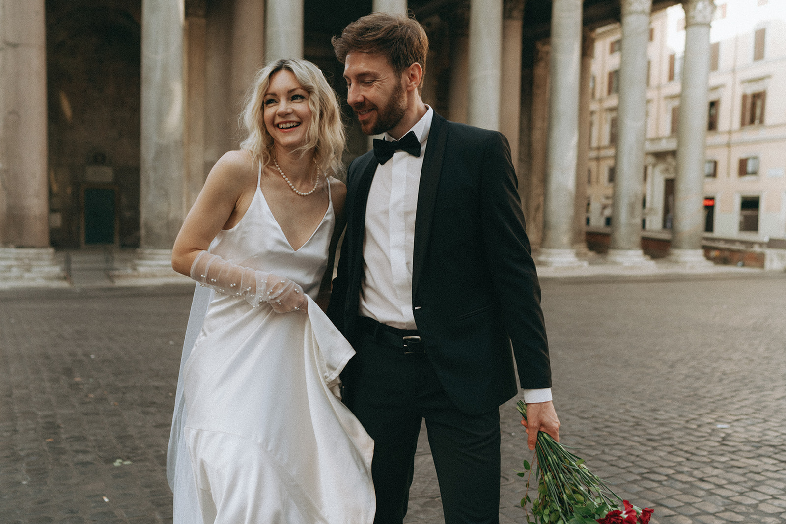 Close-up of a bride and groom in front of the Pantheon, they're close together, smiling, he is holding the bouquet of red roses by Italy elopement photographer Elsie Goodman.