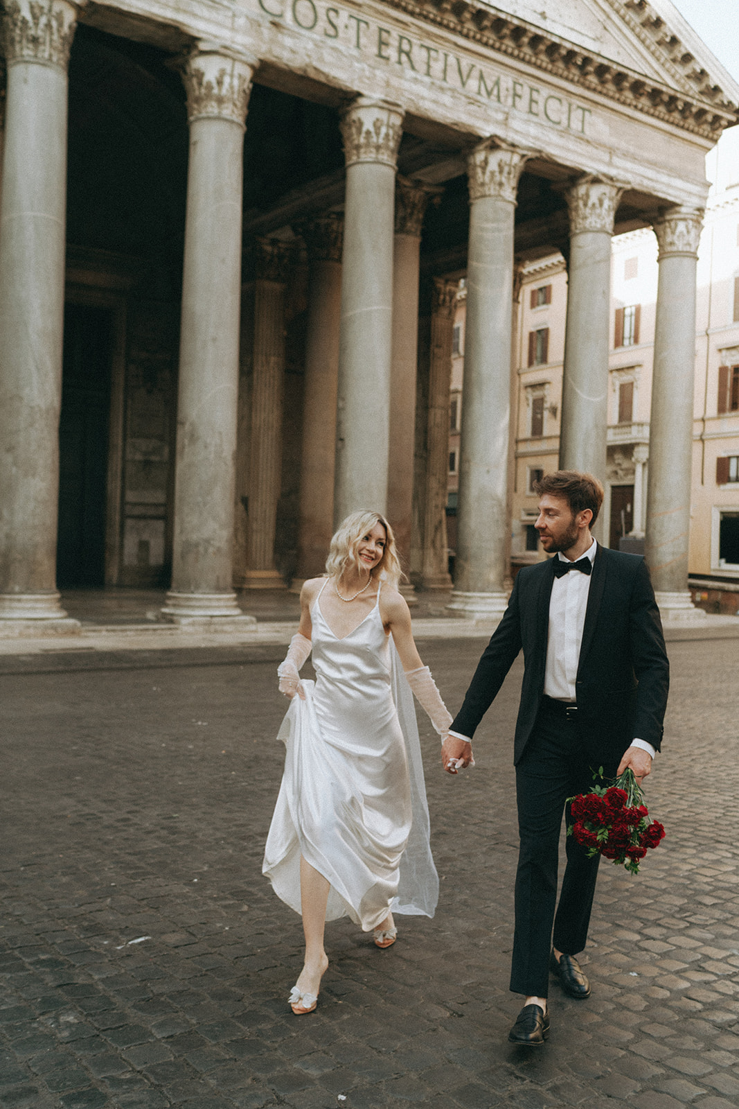 Bride and groom looking at each other, he holds the bouquet of roses as they walk in front of the Pantheon by Italy elopement photographer Elsie Goodman.