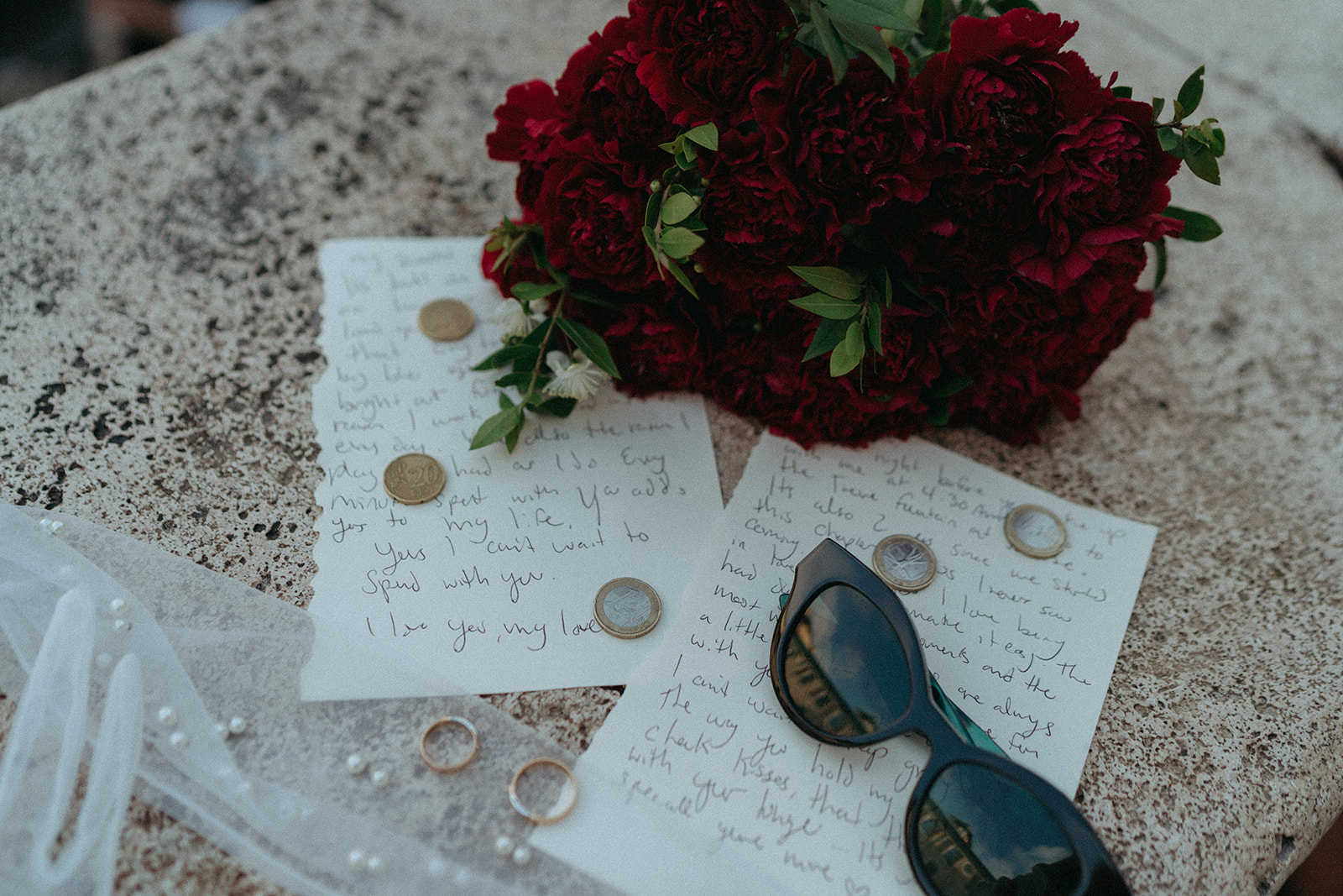 Flat lay shot of the bride and grooms vows with euros, their wedding rings, sunglasses, her gloves and her bouquet of red flowers around them by Italy elopement photographer Elsie Goodman.