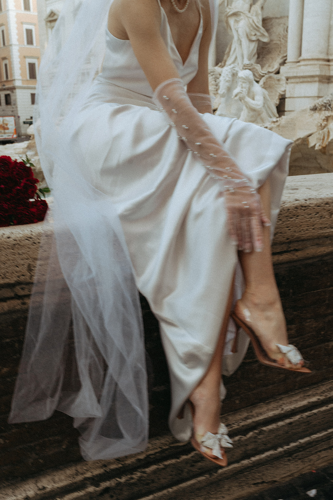 Lower shot of a bride in a long white silk dress with a long veil behind her, as she reaches down to her ankle to show off her heals with white bows on them by Italy elopement photographer Elsie Goodman.
