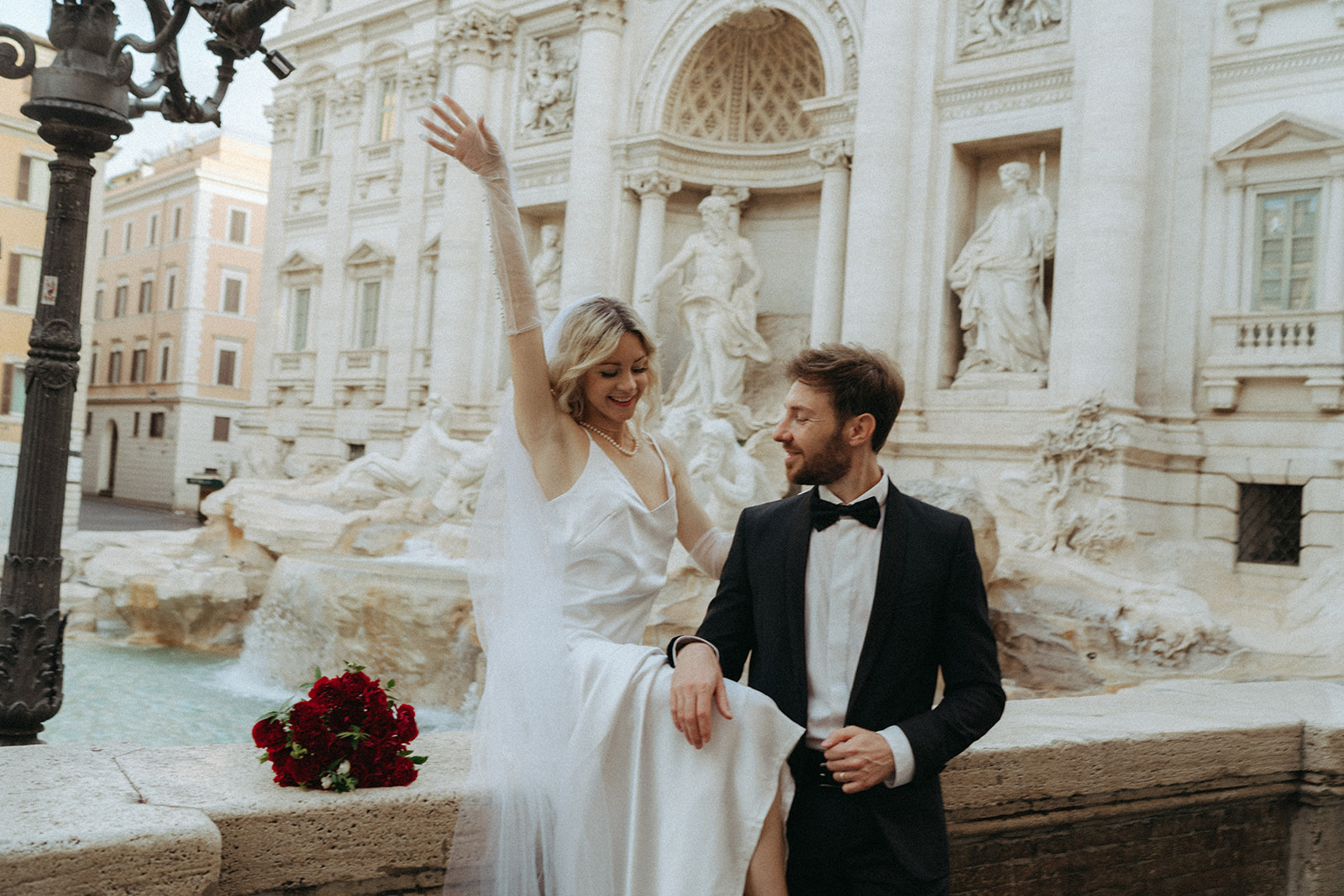 Bride sits on the border of the Trevi Fountain smiling with one hand in the air, her bouquet of red roses sits beside her. Her groom is looking at her with his hands casually on her leg by Italy elopement photographer Elsie Goodman.