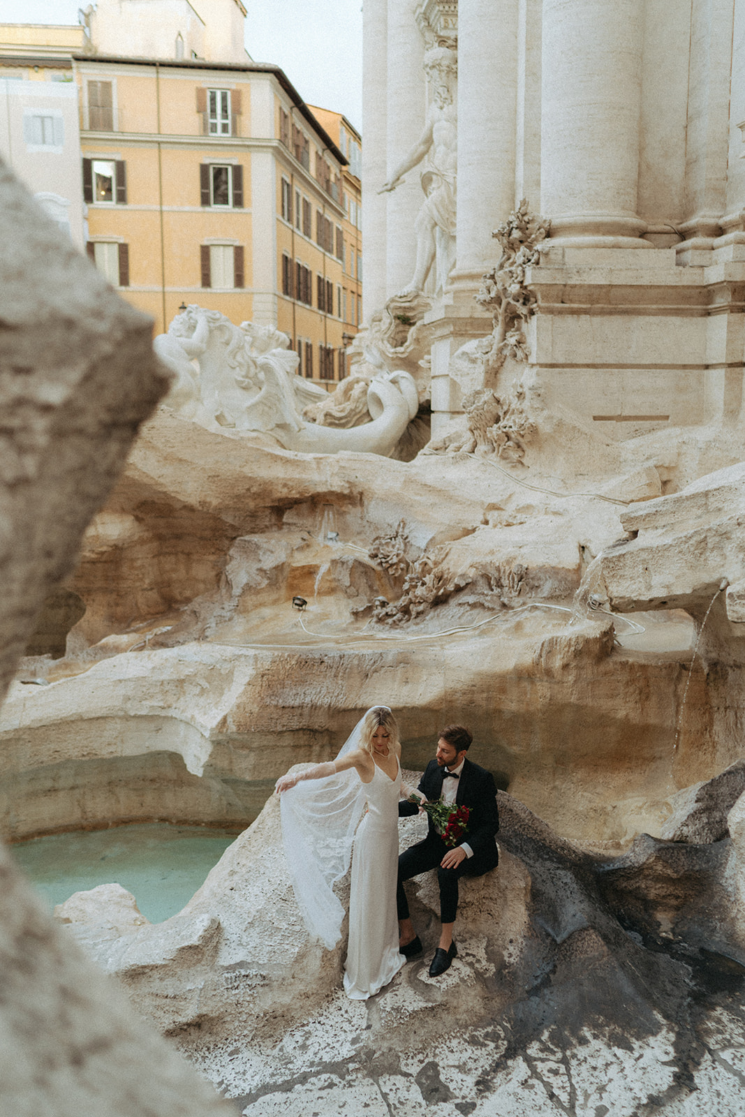 Bride holding out her veil, while the groom sits on the rough sides of the Trevi Fountain at dawn by Italy elopement photographer Elsie Goodman.
