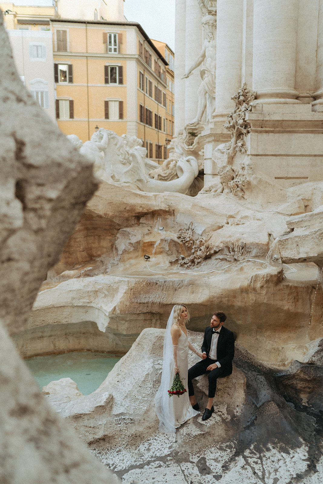 Faraway shot of the bride and groom sitting in the caves of the Trevi Fountain. The bride holds a bouquet of red roses, and groom is sitting holding her hands by Italy elopement photographer Elsie Goodman.