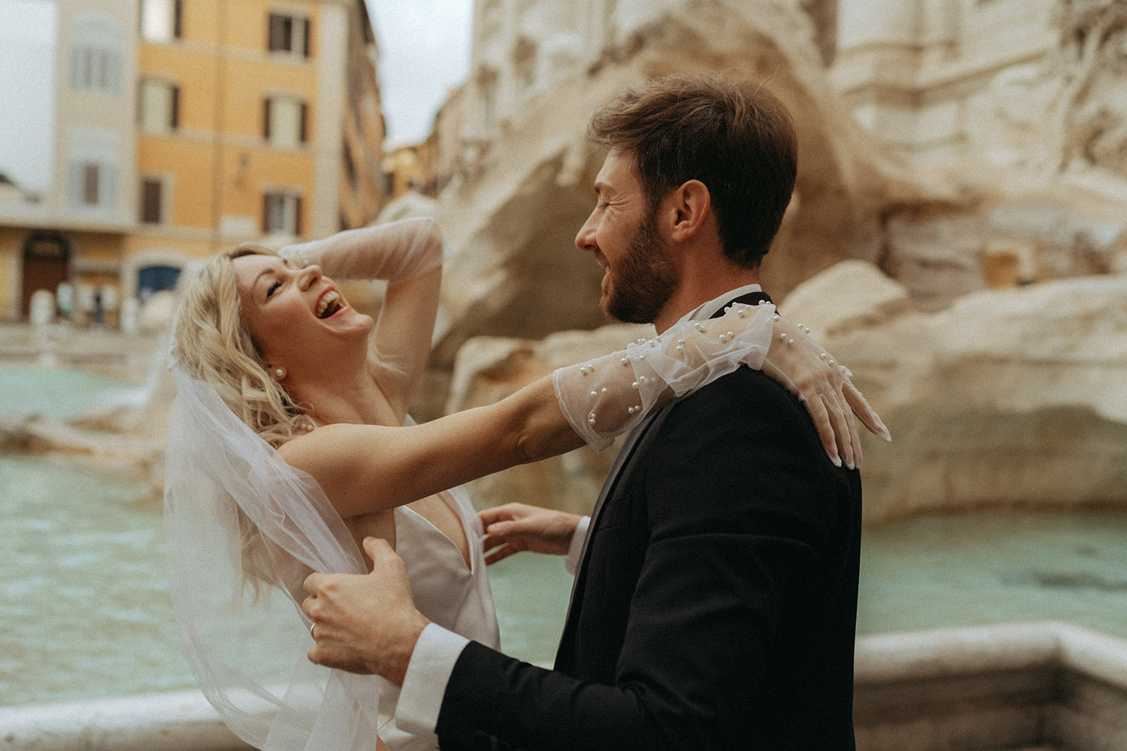 Bride holds onto her groom and leans back laughing in front of the Trevi Fountain at dawn by Italy elopement photographer Elsie Goodman.