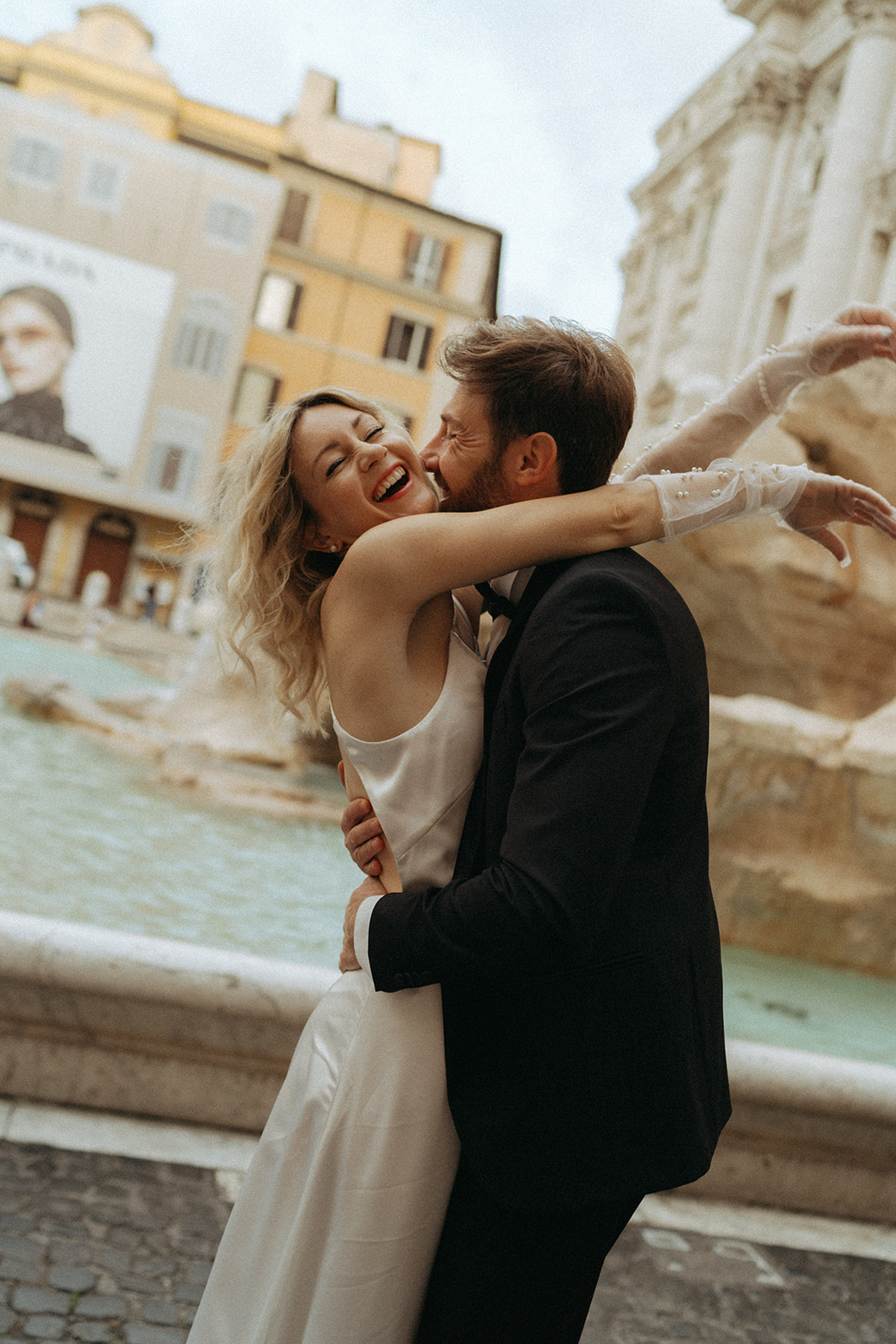 Bride and groom smiling at dawn in front of the Trevi Fountain. Her arms are around his neck, he has his arms wrapped around her leaning in for a cheek kiss by Italy elopement photographer Elsie Goodman.