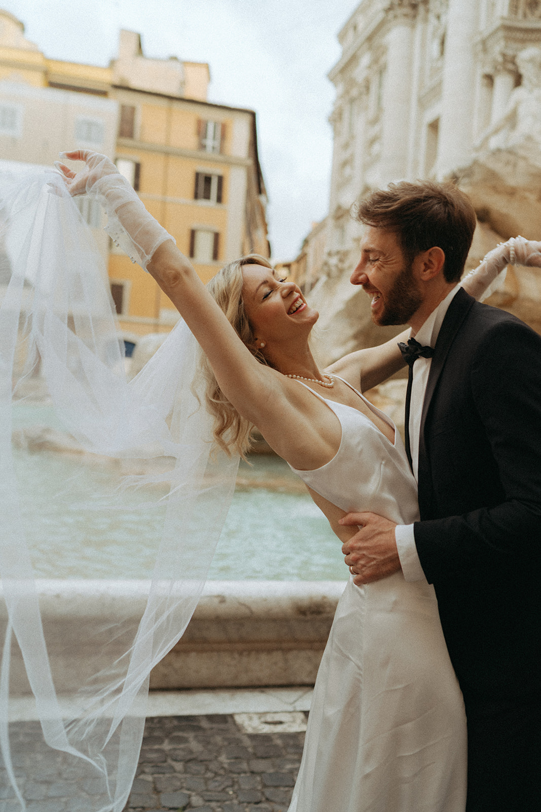 Bride throwing back her veil smiling, while the groom smiles, hands around her waist leans towards her, they are in front of the Trevi Fountain at dawn by Italy elopement photographer Elsie Goodman.