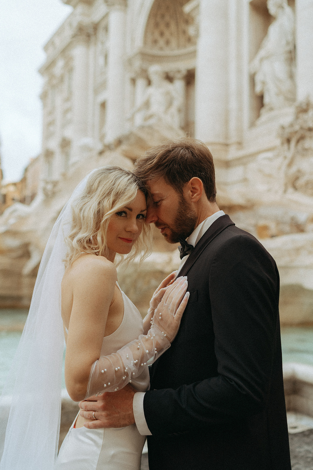 Bride and groom leaning in, the bride is looking at the camera with a slight smile in front of the Trevi Fountain at sunrise by Italy elopement photographer Elsie Goodman.