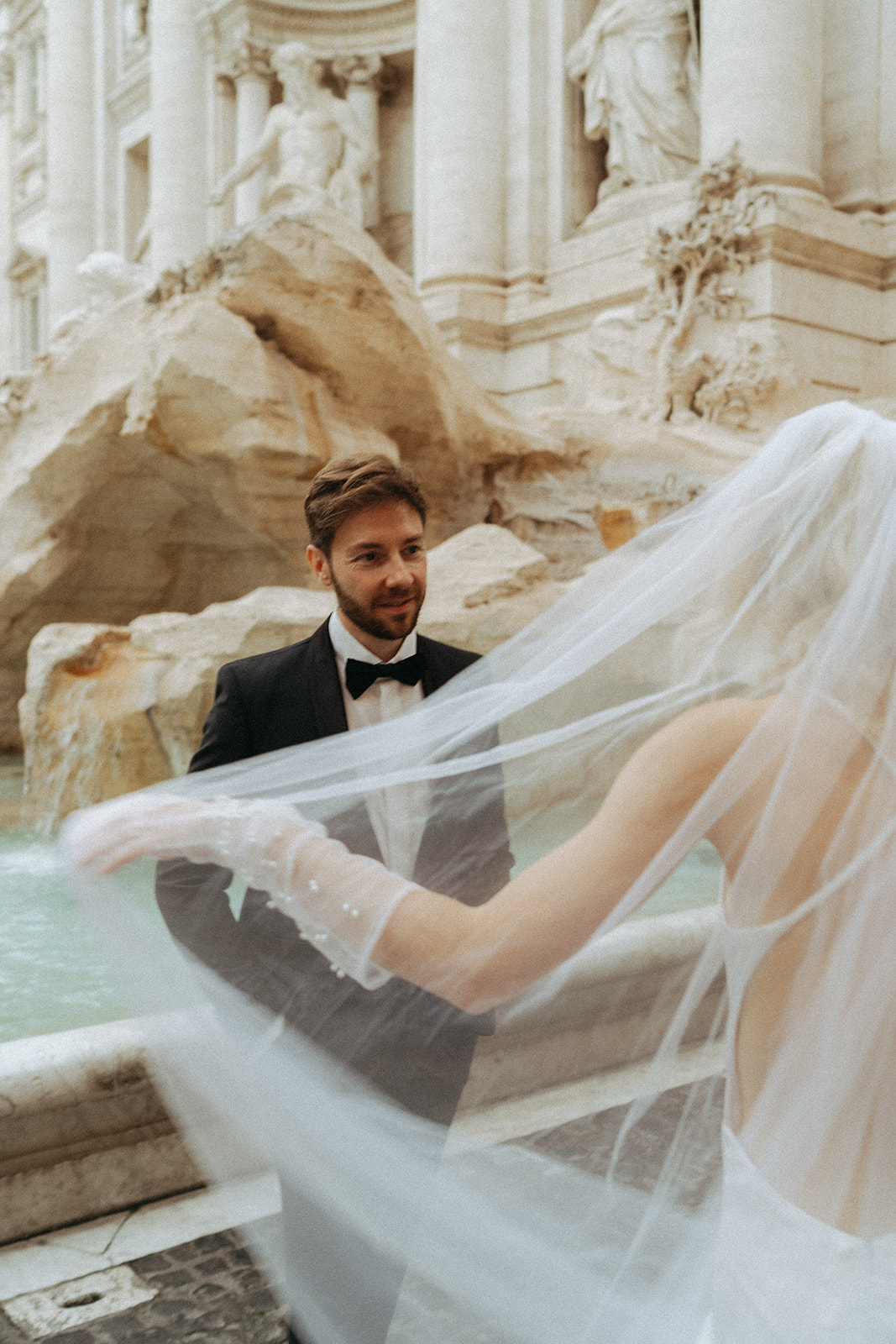 Groom looking at his bride as she throws her veil behind her. They are in front of the Trevi Fountain at sunrise by Italy elopement photographer Elsie Goodman.