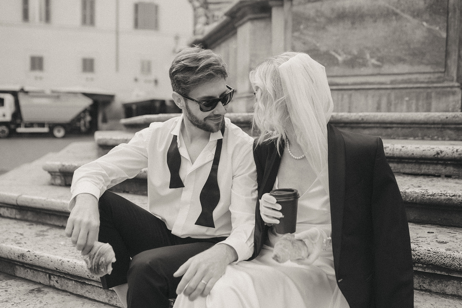 Black and white photo of a bride and groom sharing a coffee and croissant, sitting on the steps of a fountain by Rome elopement photographer Elsie Goodman.