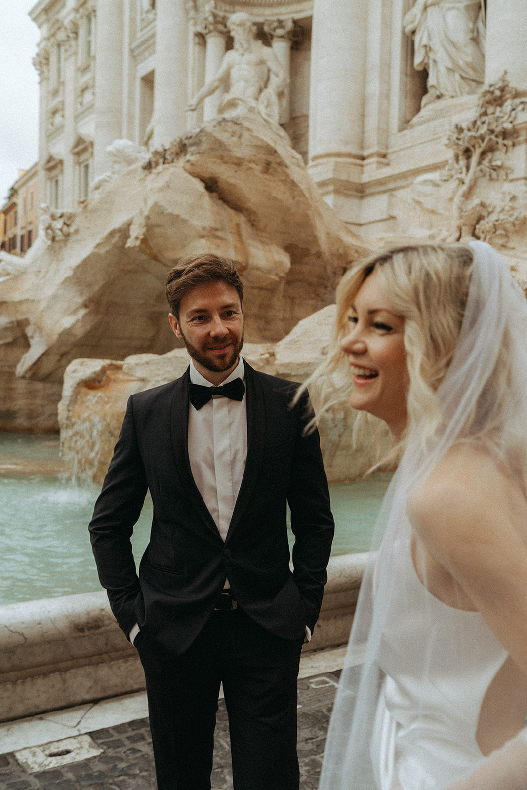 Groom looking loving at his bride as she looks away from the camera smiling in front of the Trevi Fountain at sunrise by Italy elopement photographer Elsie Goodman.