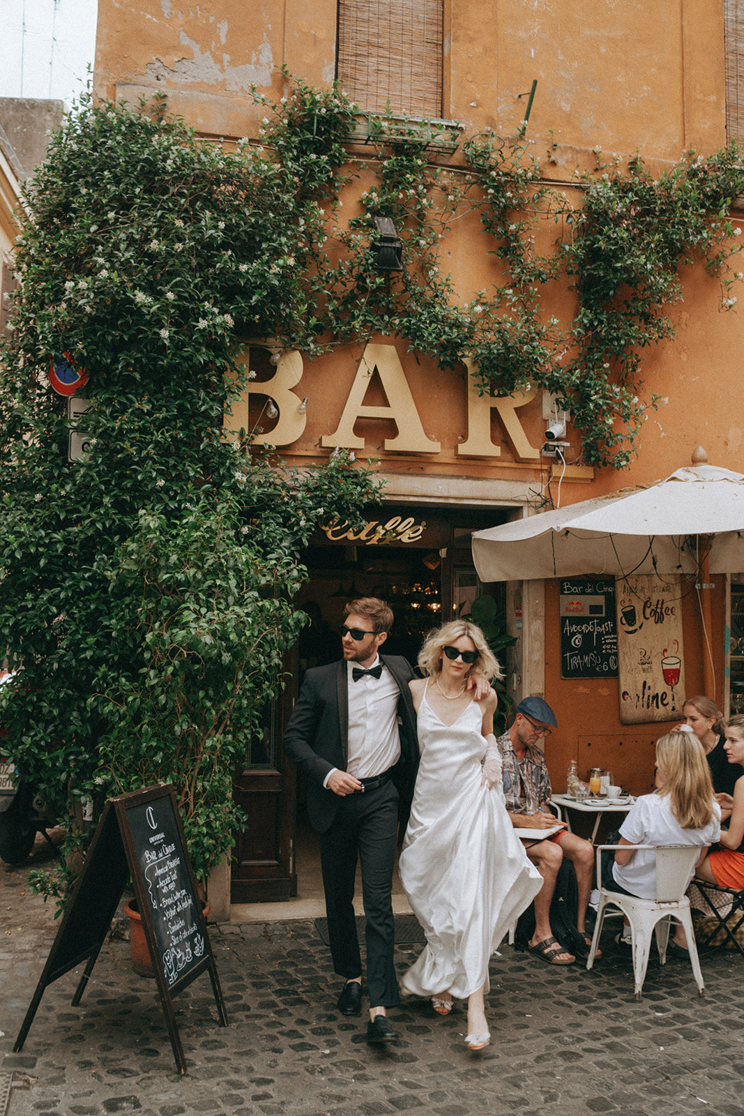 Groom with his arm around his bride, walks out of a Trastevere coffee shop by Rome elopement photographer Elsie Goodman.
