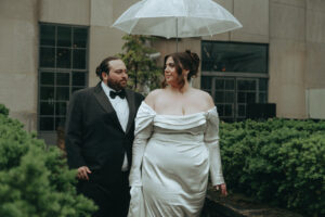 Bride and groom under a clear umbrella in the rain at their NYC elopement, groom in a tuxedo