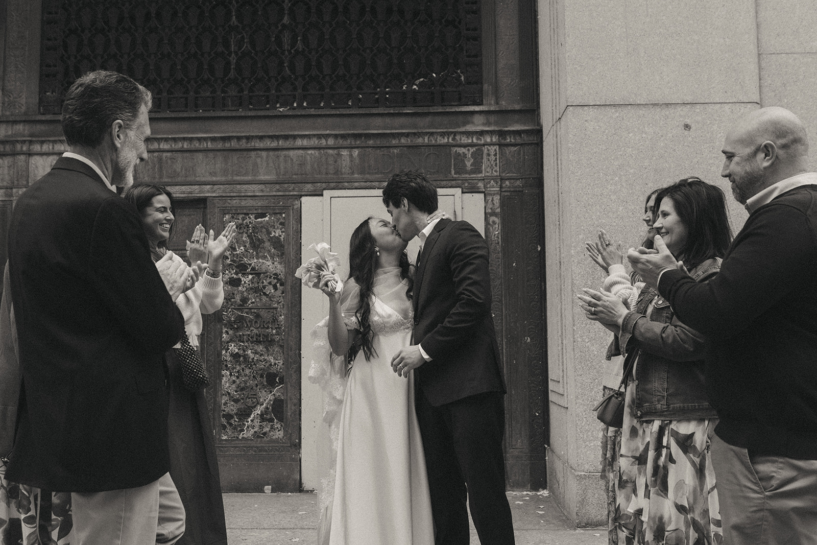 Alex + Bardo NYC Elopement Photos bride and groom kiss when coming out of nyc city hall after getting married with loved ones clapping, shot by elsie goodman, a destination, wedding and NYC elopement photographer