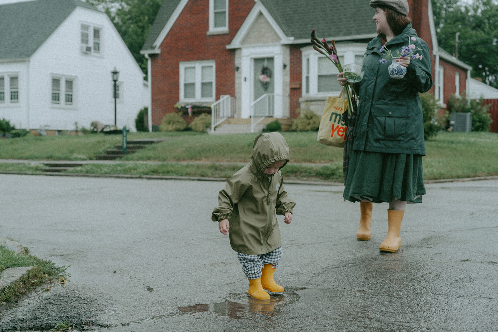 child in rain jacket and yellow rain boots playing in a puddle as mom walks behind with flowers and vases in her hands by maternity, family, and wedding photographer Elsie Goodman.