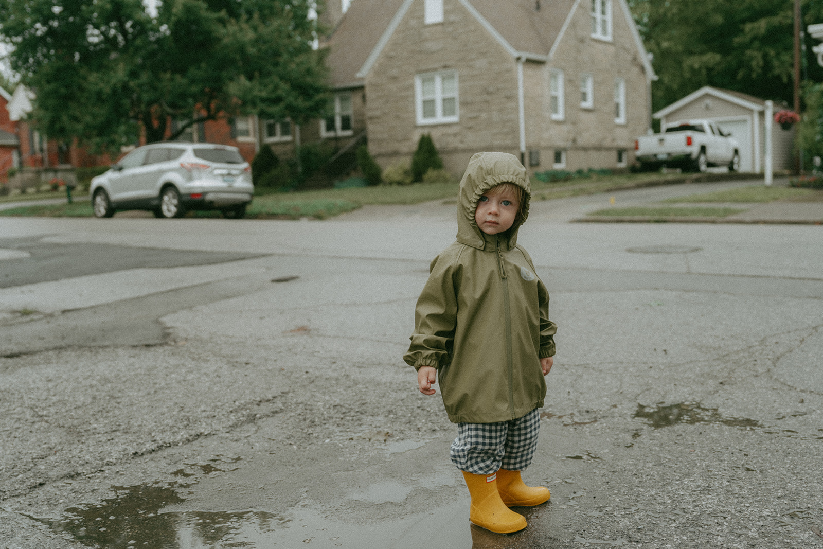 Child looking at the camera in a green rain jacket, black and white checkered pants and yellow rain boots by maternity, family, and wedding photographer Elsie Goodman.