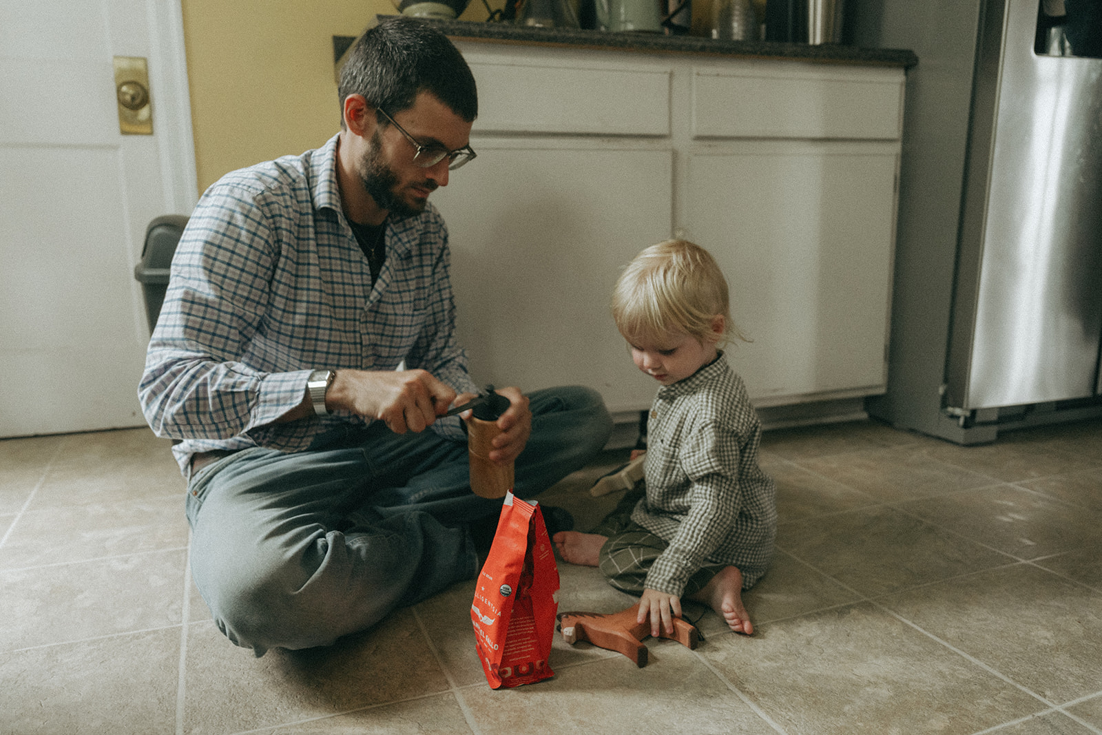 Dad and child sitting on the kitchen floor grinding some coffee by maternity, family, and wedding photographer Elsie Goodman.