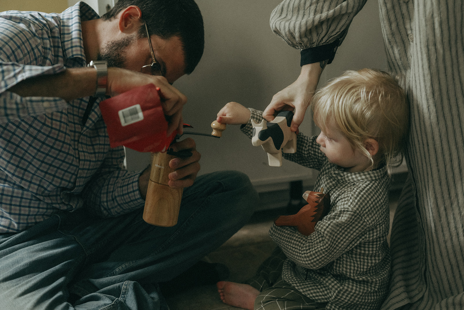 Dad pouring coffee beans into a hand cranked coffee grinder while his child helps, Mom is leaning over holding a wooden toy cow by maternity, family, and wedding photographer Elsie Goodman.