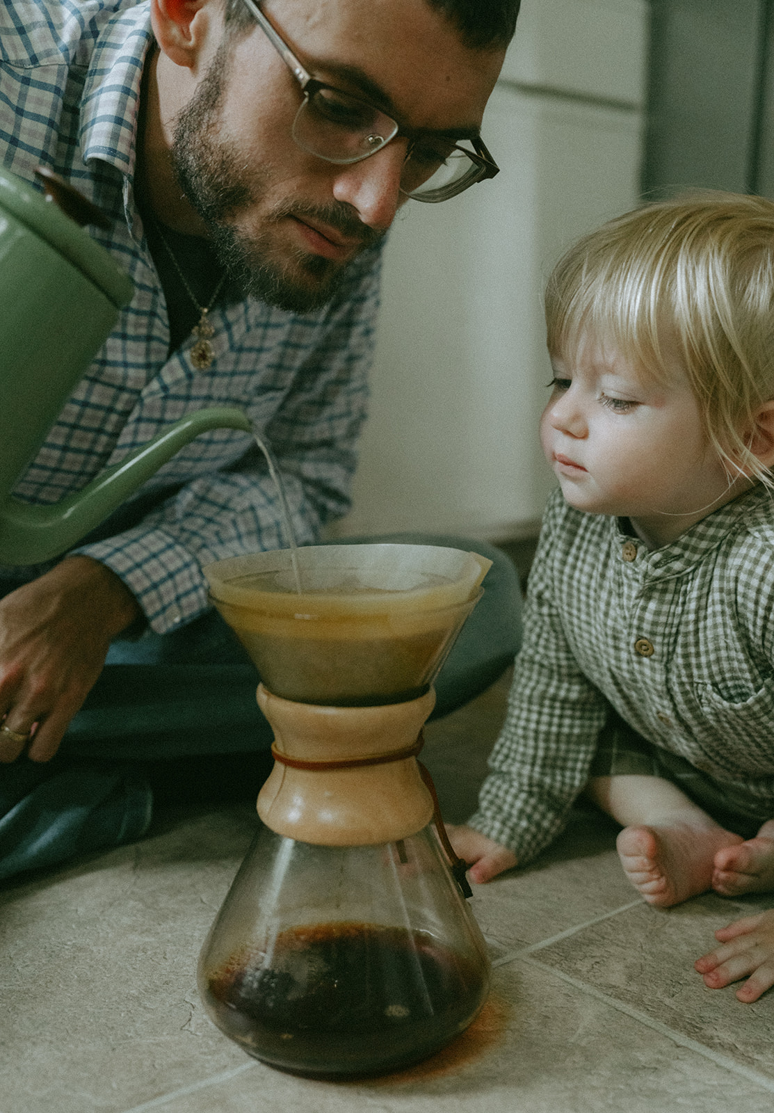 Dad sitting on the kitchen floor with his child as he pours water into a Chemex, they are both leaning in close by maternity, family, and wedding photographer Elsie Goodman.