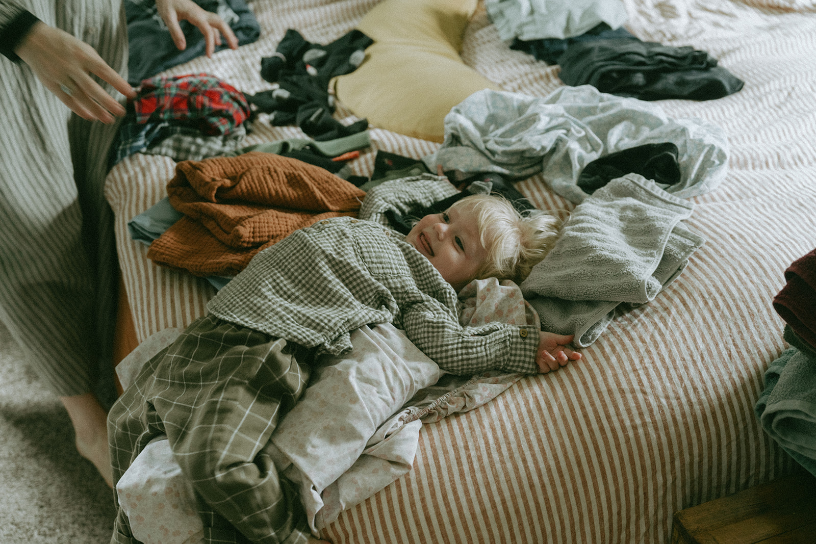 Child laying on top of clothes being folded on the bed by maternity, family, and wedding photographer Elsie Goodman.