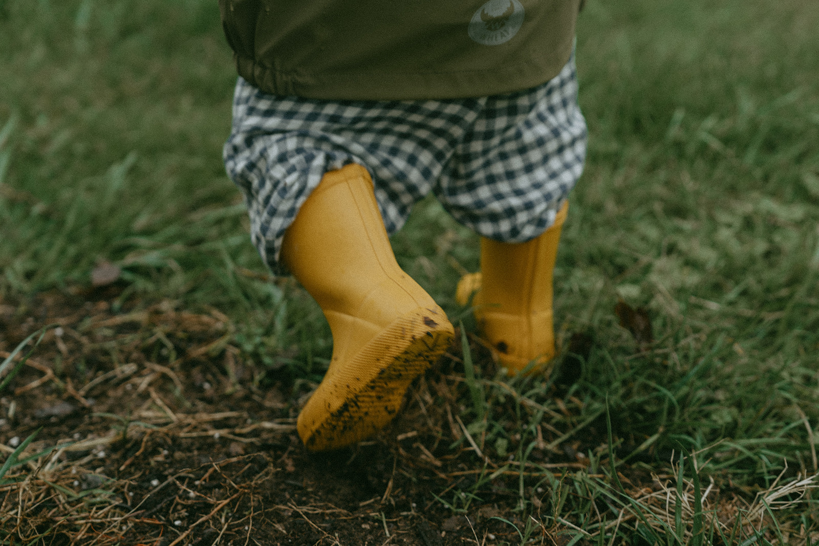 Kid walking away in yellow rain boots in the grass and mud by maternity, family, and wedding photographer Elsie Goodman.