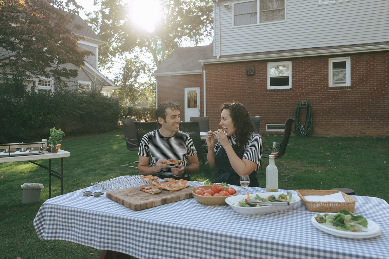Man and woman sitting at a picnic table in the backyard eating pizza they just made by the wedding and couples photographer, Elsie Goodman.