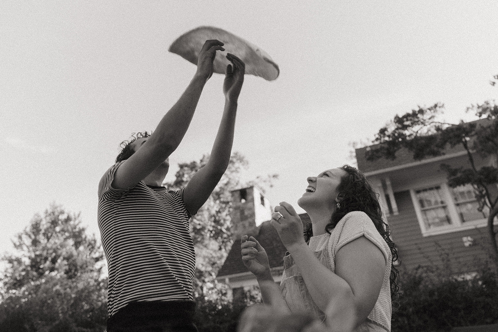 Black and white photo of a man throwing dough up in the air, while the woman laughs looking up by the wedding and couples photographer, Elsie Goodman.