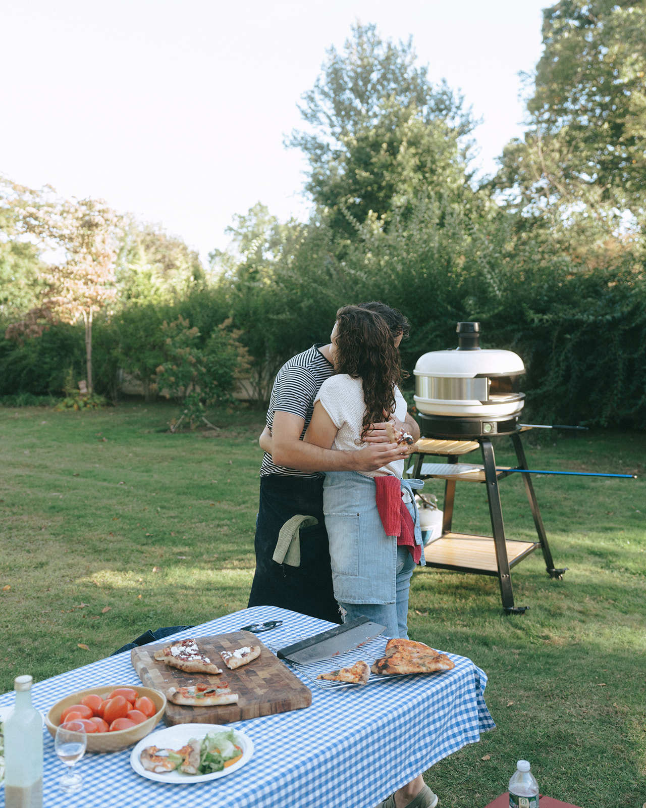 Couple hugging outside with pizza making ingredients and picnic table around them by wedding and couples photographer Elsie Goodman.