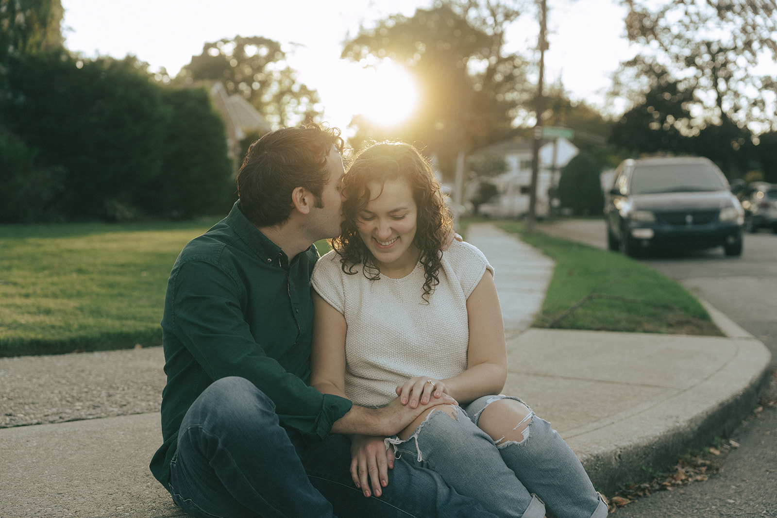 Couple sitting on a street curb, the man is kissing the side of the woman's face, taken at sunset by wedding and couples photographer Elsie Goodman.