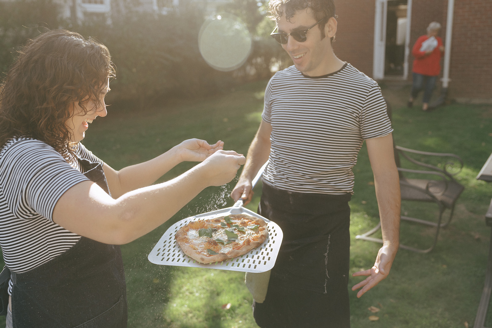 Couple making pizza in the backyard, the man is holding a pizza on a peel, the woman is sprinkling parmesan by couples photographer, Elsie Goodman.